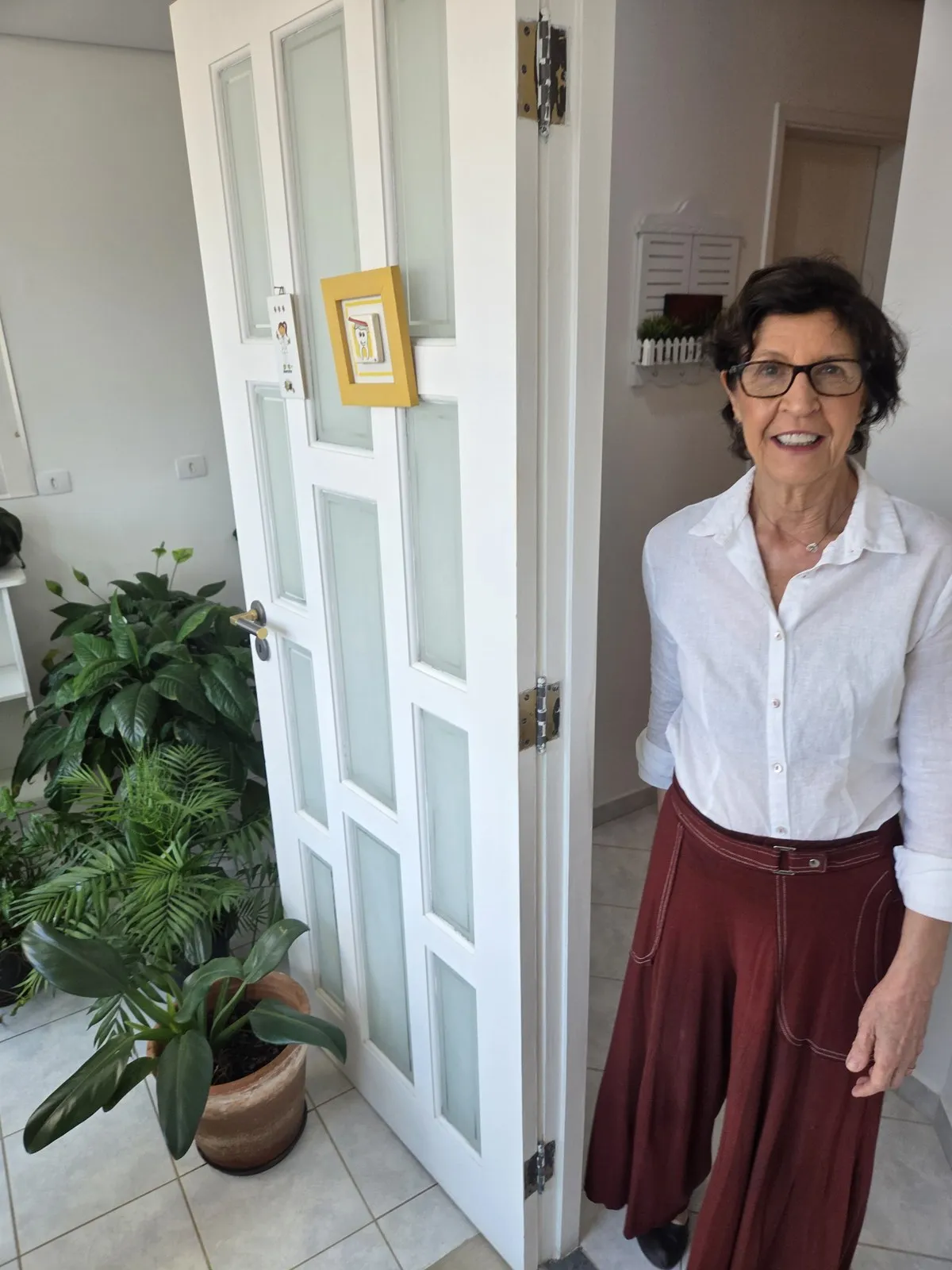Ana Junqueira no consultório, ao lado da porta de entrada, com luz natural.